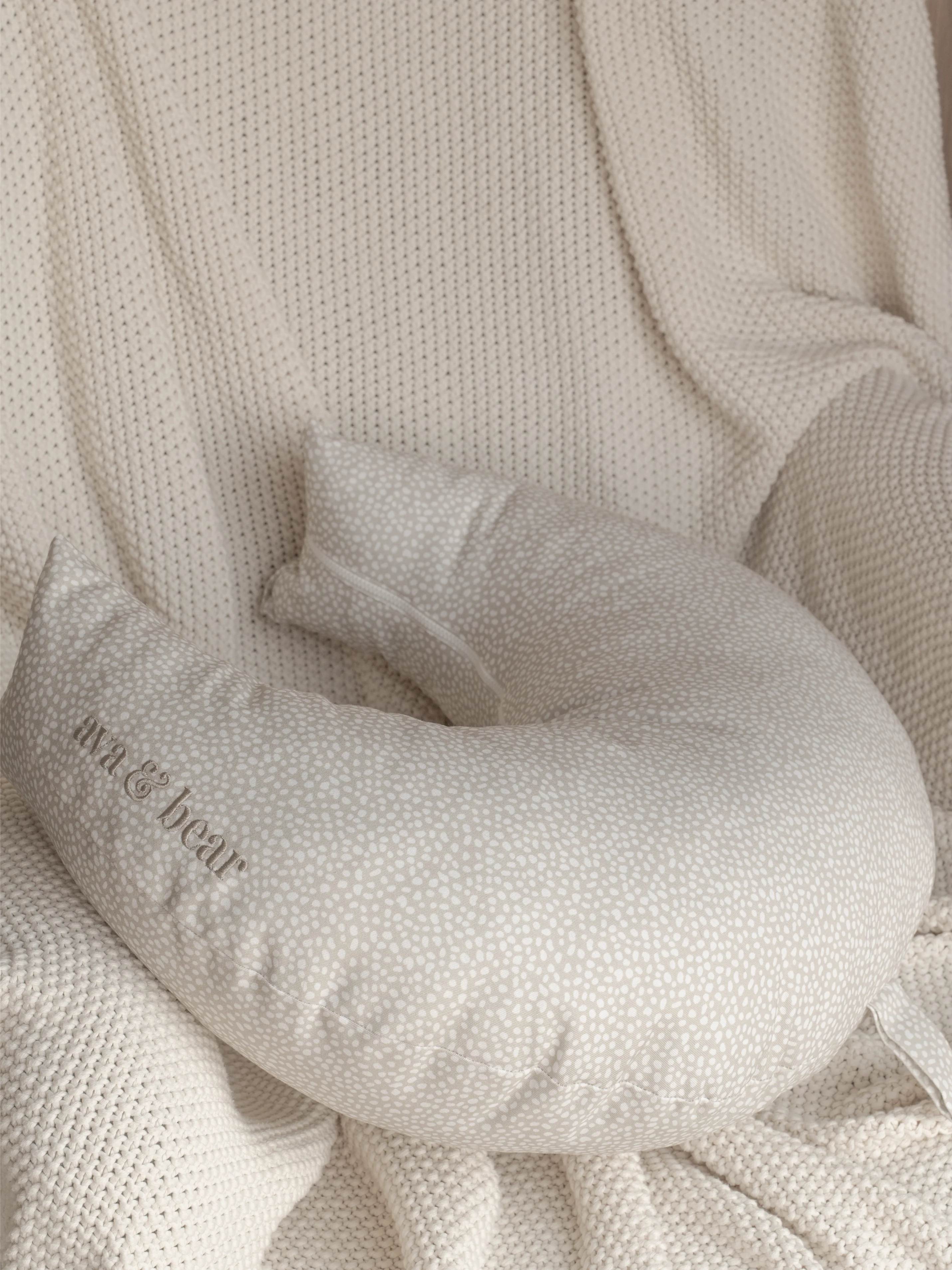 Crescent moon-shaped Ava & Bear nursing cushion in stone spot design, crafted from 100% cotton twill for durability and comfort, featuring a convenient loop handle for easy portability, ideal for supporting mother and baby during feeding.